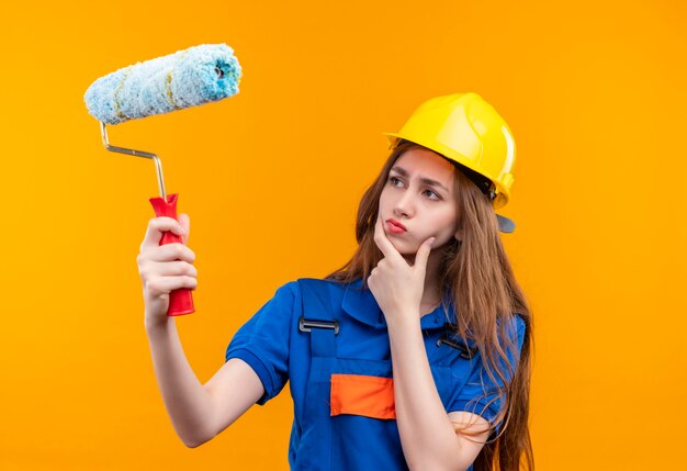 Operaio costruttore di giovane donna in uniforme da costruzione e casco di sicurezza che tiene il rullo di vernice e pennello guardando il rullo con espressione scettica, pensando in piedi sopra la parete arancione