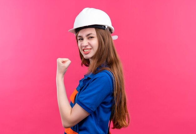 Operaio del costruttore della giovane donna in uniforme da costruzione e casco di sicurezza che alza il pugno che sembra sicuro, in posa come un vincitore in piedi sopra il muro rosa