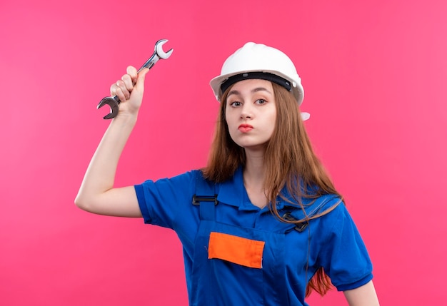 Operaio del costruttore della giovane donna in uniforme da costruzione e casco di sicurezza che oscilla una chiave con la faccia seria che sta sopra il muro rosa