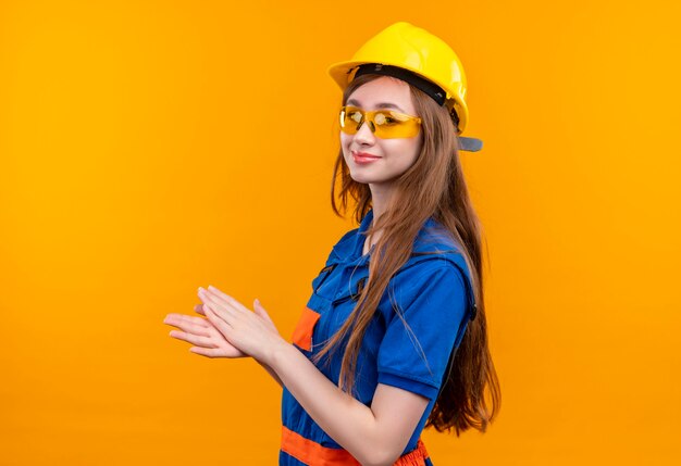 Operaio del costruttore della giovane donna in uniforme da costruzione e casco di sicurezza che sembra applaudire fiducioso in piedi
