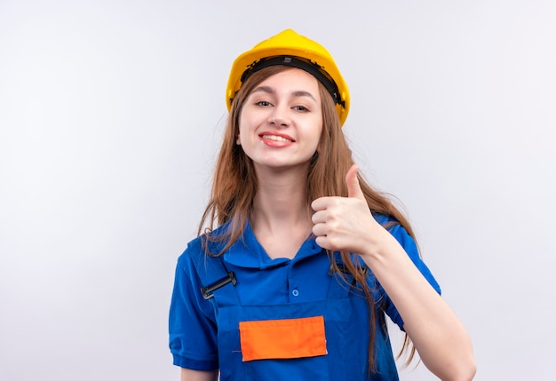 Operaio del costruttore della giovane donna in uniforme da costruzione e casco di sicurezza che sorride allegramente mostrando i pollici in su in piedi sopra il muro bianco