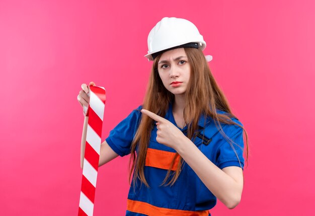 Operaio del costruttore della giovane donna in uniforme da costruzione e casco di sicurezza che tiene il nastro adesivo che indica con il dito indice ad esso con la faccia seria che sta sopra il muro rosa