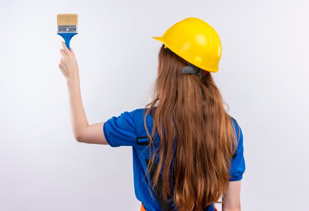 Operaio del costruttore della giovane donna in uniforme da costruzione e casco di sicurezza in piedi con la schiena che tiene il pennello sul muro bianco