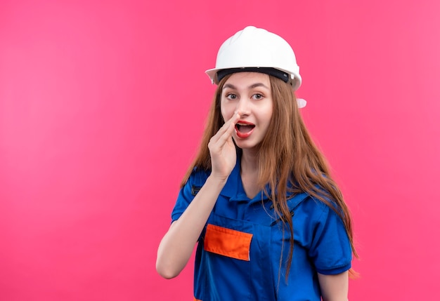 Operaio del costruttore della giovane donna in uniforme della costruzione e casco di sicurezza che dice un segreto con la mano vicino alla bocca che sta sopra la parete rosa