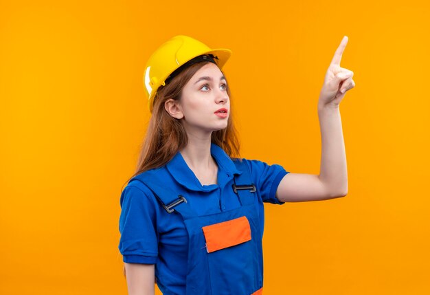 Operaio del costruttore della giovane donna in uniforme della costruzione e casco di sicurezza che indica con il dito in su che sembra fiducioso in piedi sopra la parete arancione