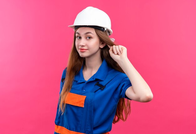 Operaio del costruttore della giovane donna in uniforme della costruzione e casco di sicurezza che solleva il pugno che sembra sicuro, concetto del vincitore che sta sopra la parete rosa