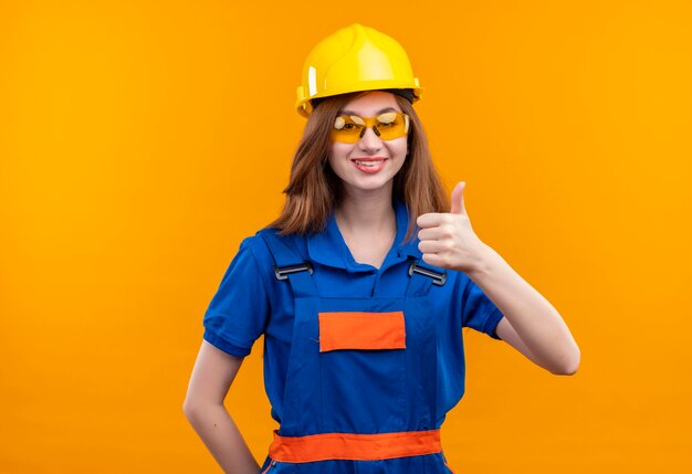 Operaio del costruttore della giovane donna in uniforme della costruzione e casco di sicurezza che sorride ampiamente che mostra i pollici in su che si leva in piedi sopra la parete arancione