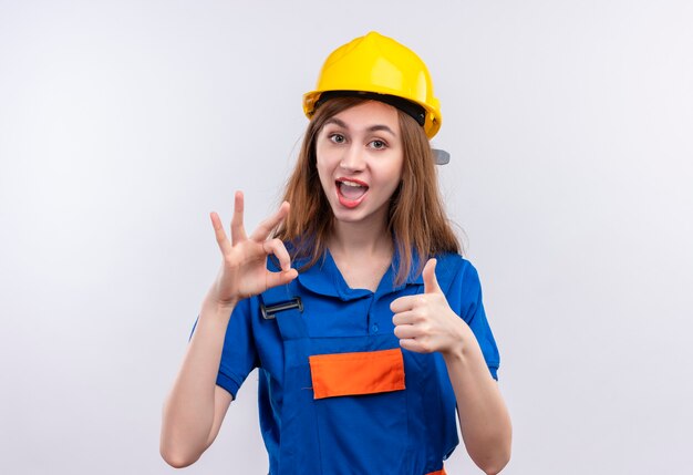 Operaio del costruttore della giovane donna in uniforme della costruzione e casco di sicurezza che sorride ampiamente che mostra i pollici in su e segno giusto che sta sopra il muro bianco