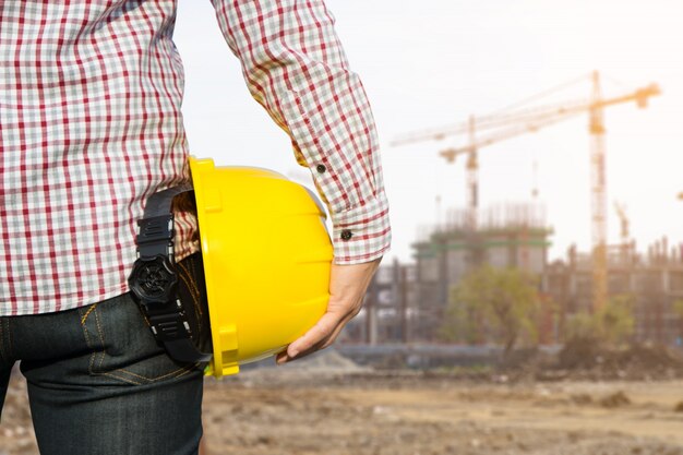 Operaio di ingegnere della mano che tiene casco di sicurezza giallo con la costruzione sullo sfondo del luogo.