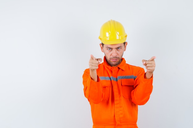 Operaio edile che indica in uniforme, casco e che sembra fiducioso. vista frontale.