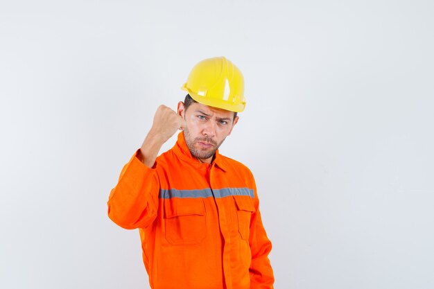 Operaio edile che mostra il pugno chiuso in uniforme, casco e guardando fiducioso, vista frontale.