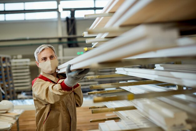 Operaio manuale con maschera facciale che impila assi di legno su uno scaffale in un'officina di falegnameria