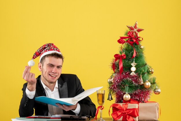Operaio maschio di vista frontale che tiene i documenti intorno al piccolo albero di natale e presenta sul lavoro giallo di Natale di colore dell'ufficio del nuovo anno dello scrittorio