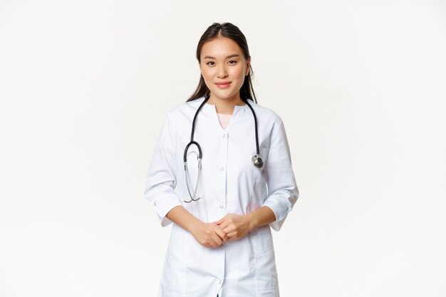 Operaio medico asiatico sorridente con lo stetoscopio, indossando l'uniforme del medico, guardando utile al paziente, in piedi su sfondo bianco.