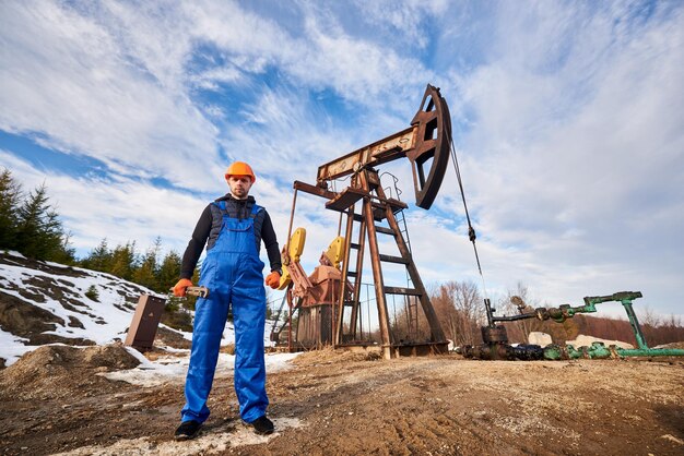 Operaio petrolifero in uniforme e casco che lavora in un giacimento petrolifero accanto a un martinetto della pompa in una giornata di sole