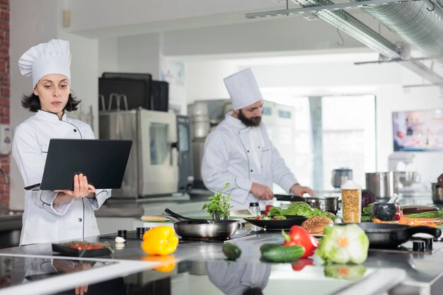 Operaio professionista dell'industria alimentare con computer alla ricerca di pasti gastronomici. Capo chef donna con laptop brainstorming guarnire idee per la ricetta di un piatto gourmet mentre si è in piedi nella cucina del ristorante.