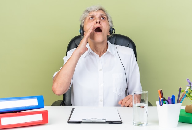 Operatore di call center femminile caucasica scioccato sulle cuffie seduto alla scrivania con strumenti da ufficio con la mano vicino alla bocca che guarda in alto