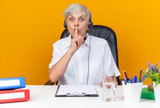 Operatore di call center femminile caucasico ansioso sulle cuffie seduto alla scrivania con strumenti da ufficio facendo gesto di silenzio isolato sulla parete arancione