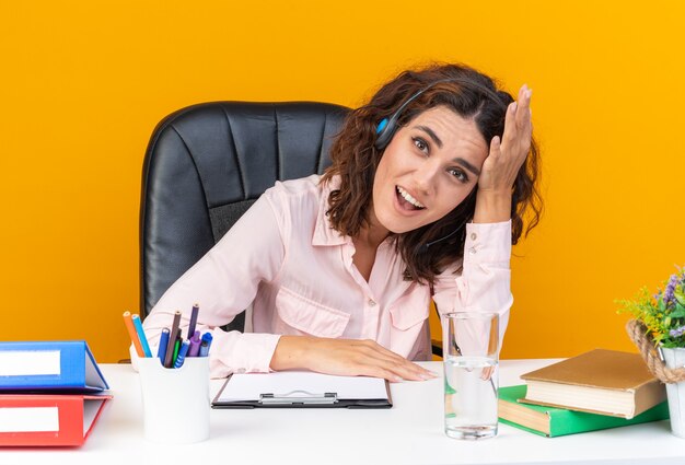 Operatore di call center femminile piuttosto caucasico sorpreso sulle cuffie seduto alla scrivania con strumenti da ufficio mettendo la mano sulla fronte guardando davanti