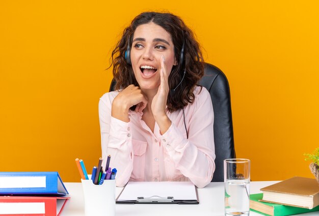 Operatore di call center femminile piuttosto caucasico sorpreso sulle cuffie seduto alla scrivania con strumenti da ufficio tenendo la mano vicino alla bocca guardando il lato isolato sul muro arancione