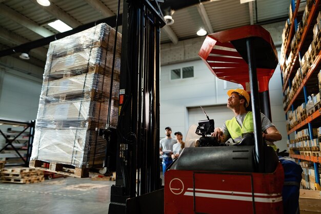 Operatore di carrelli elevatori che carica merci mentre lavora in un magazzino I suoi colleghi sono in background