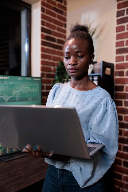 Operatore professionale di società di hedge fund con laptop durante la revisione delle statistiche finanziarie in tempo reale nell'area di lavoro dell'ufficio. Agente del mercato azionario Forex in piedi davanti a una workstation multi monitor.