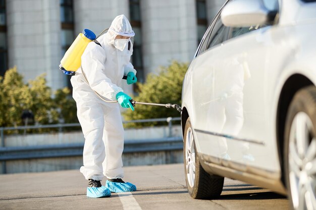 Operatore sanitario in tuta protettiva che spruzza pneumatici per auto durante la disinfezione della città a causa della pandemia COVID19