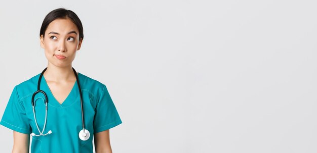 Operatori sanitari, prevenzione del virus, concetto di campagna di quarantena. Premurosa dottoressa asiatica, infermiera in camice che guarda nell'angolo in alto a sinistra e sorride incerta, pensando, sfondo bianco.