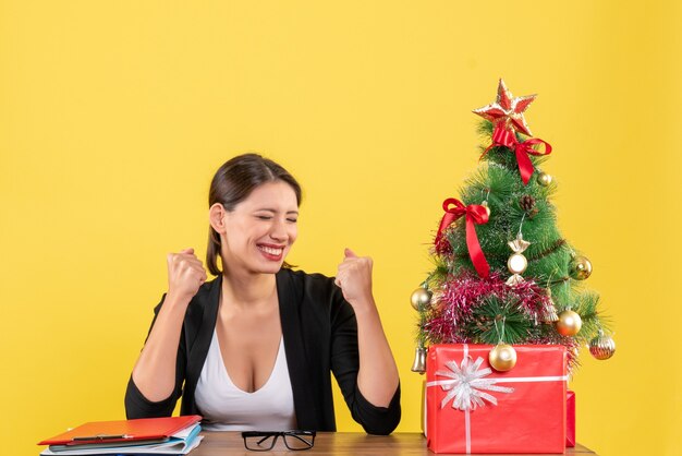 Orgogliosa giovane donna in tuta con albero di Natale decorato in ufficio su giallo