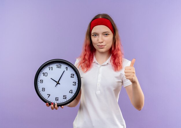 Orologio da parete della holding della giovane donna sportiva che mostra i pollici in su sorridente che sta sopra la parete viola