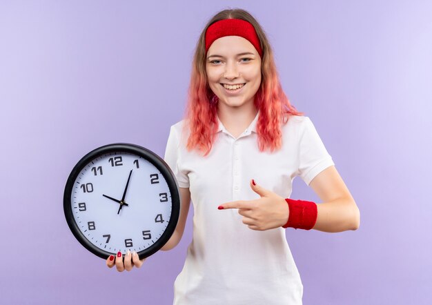 Orologio da parete della tenuta della giovane donna sportiva che indica con il dito che sorride allegramente in piedi sopra la parete viola