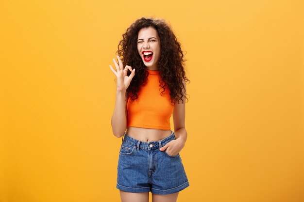 Ottima festa piace. Gioiosa entusiasta giovane femmina dai capelli ricci con rossetto rosso in elegante top ritagliato strizzando l'occhio eccitato sorridendo e mostrando segno giusto o perfetto in posa su sfondo arancione.