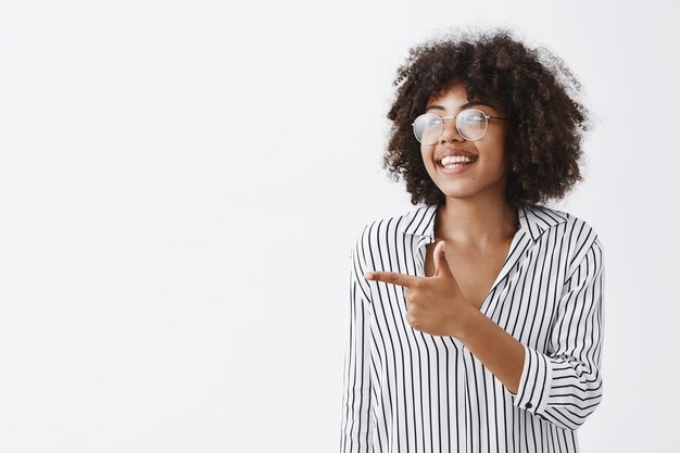 ottimista amichevole e gioiosa donna dalla pelle scura in camicetta e occhiali che guarda e indica felicemente a destra sorridendo ampiamente