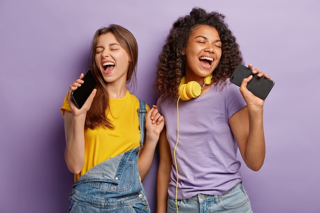 Ottimiste e compiaciute donne di razza mista cantano la canzone preferita negli smartphone, si divertono e godono della musica, tengono gli occhi chiusi, si muovono attivamente