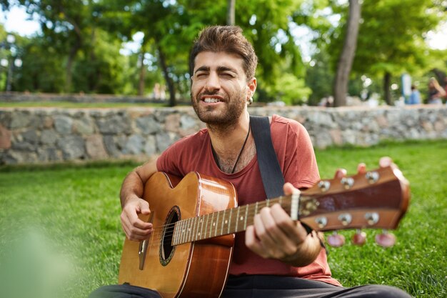 Outdoor ritratto di bel ragazzo romantico seduto sull'erba nel parco e suonare la chitarra