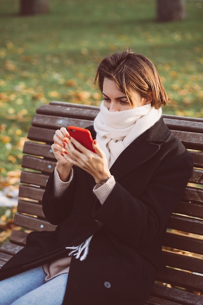 Outdoor ritratto di donna caucasica che indossa cappotto invernale nero e sciarpa seduto sulla panchina nel parco con il telefono cellulare.