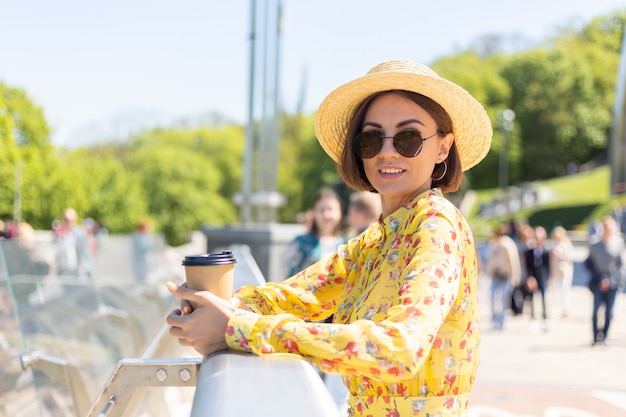 Outdoor ritratto di donna in abito estivo giallo e cappello con una tazza di caffè, godersi il sole, si trova sul ponte con vista mozzafiato sulla città