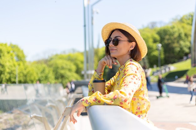 Outdoor ritratto di donna in abito estivo giallo e cappello con una tazza di caffè, godersi il sole, si trova sul ponte con vista mozzafiato sulla città