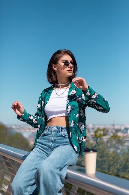 Outdoor ritratto di donna in camicia verde con una tazza di caffè che gode del sole, si trova sul ponte con vista mozzafiato sulla città al mattino