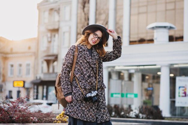 Outdoor ritratto di elegante giovane donna con zaino marrone che indossa cappotto e cappello. Donna attraente con i capelli ricci che parla sul telefono mentre beve il caffè per strada