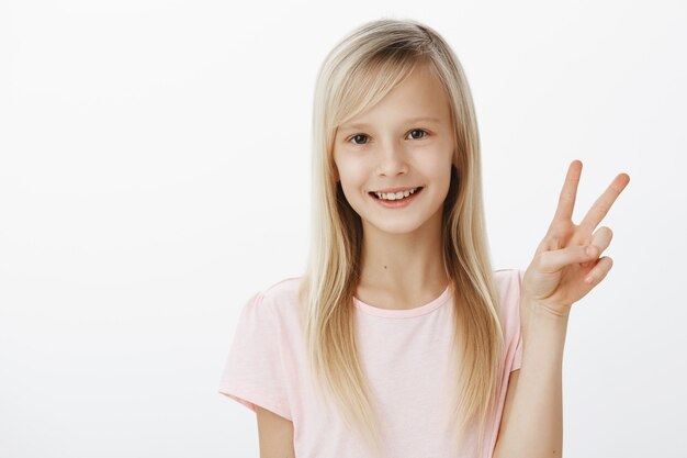 Pace a tutti i miei fan. Ritratto di adorabile ragazzina europea alla moda in maglietta rosa che mostra il gesto di vittoria e sorride ampiamente, sentendosi spensierata e fiduciosa sul muro grigio