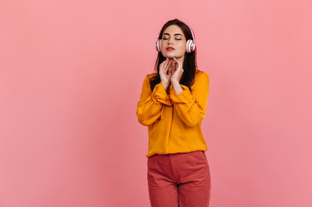 Pacificata signora dai capelli scuri in camicia gialla e pantaloni luminosi gode di musica classica in cuffia sul muro rosa.