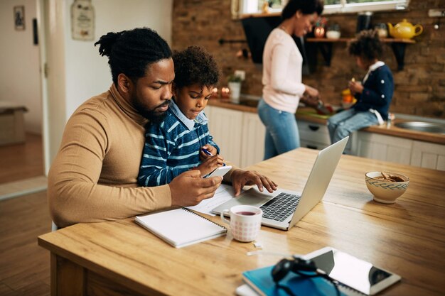 Padre afroamericano che usa lo smartphone mentre lavora al computer portatile a casa