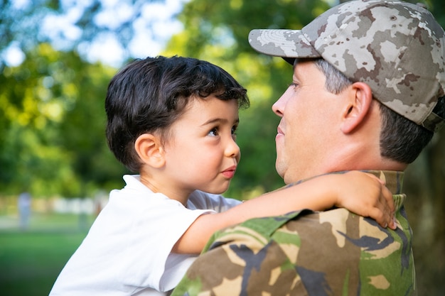 Padre allegro che tiene il piccolo figlio in braccio, abbracciando il ragazzo all'aperto dopo il ritorno dal viaggio di missione militare. Colpo del primo piano. Ricongiungimento familiare o concetto di ritorno a casa