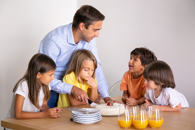 Padre caucasico che taglia deliziosa torta per i bambini. Simpatici ragazzini che circondano il tavolo, festeggiano il compleanno insieme, parlano e aspettano il dessert. Infanzia, celebrazione e concetto di vacanza