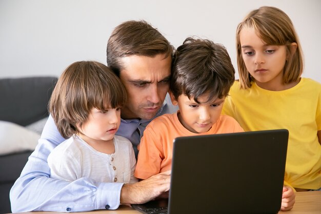 Padre concentrato e bambini seri che guardano lo schermo. Papà di mezza età caucasico che scrive sullo schermo del laptop e bambini che guardano il suo lavoro. Concetto di paternità, infanzia e tecnologia digitale