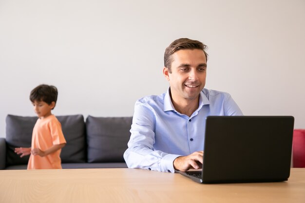 Padre felice che naviga in Internet quando il ragazzo gioca vicino a lui. Papà caucasico utilizzando il computer portatile e lavorando a casa.