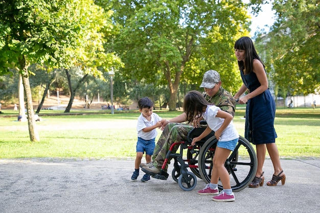 Padre militare disabile parlando con bambini carini. Papà di mezza età caucasico in uniforme mimetica all'aperto con la famiglia. Mamma graziosa che spinge la sedia a rotelle. Ricongiungimento familiare e veterano del concetto di guerra