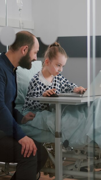 Padre seduto con la figlia malata nel reparto ospedaliero mentre gioca ai videogiochi di terapia online usando il laptop