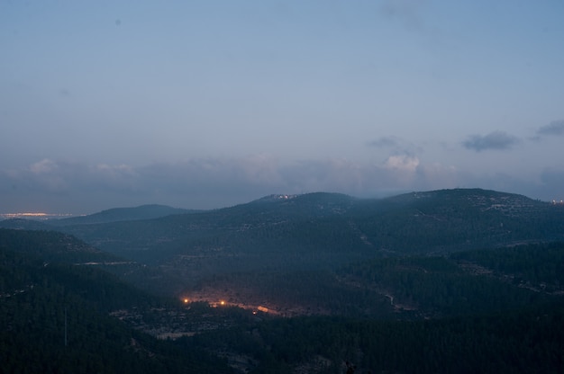 Paesaggio di colline ricoperte di boschi e luci sotto un cielo nuvoloso durante la sera